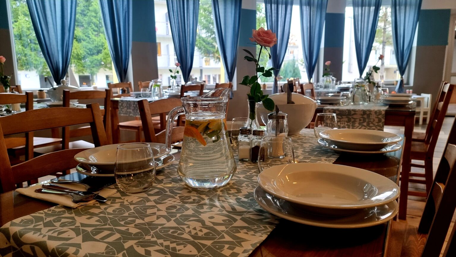 Nakryty stół w bistro Baltic Resort z karafką wody z cytrusami, talerzami i różą w wazonie, w tle okna z niebieskimi zasłonami