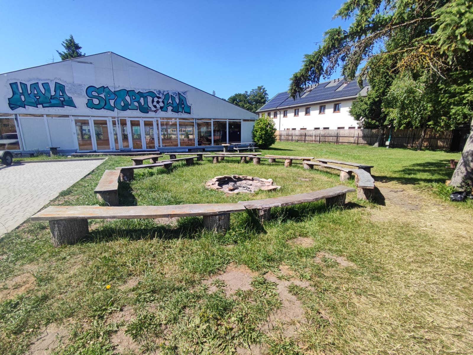 Palenisko otoczone drewnianymi ławami na terenie ośrodka Baltic Resort, w tle hala sportowa
